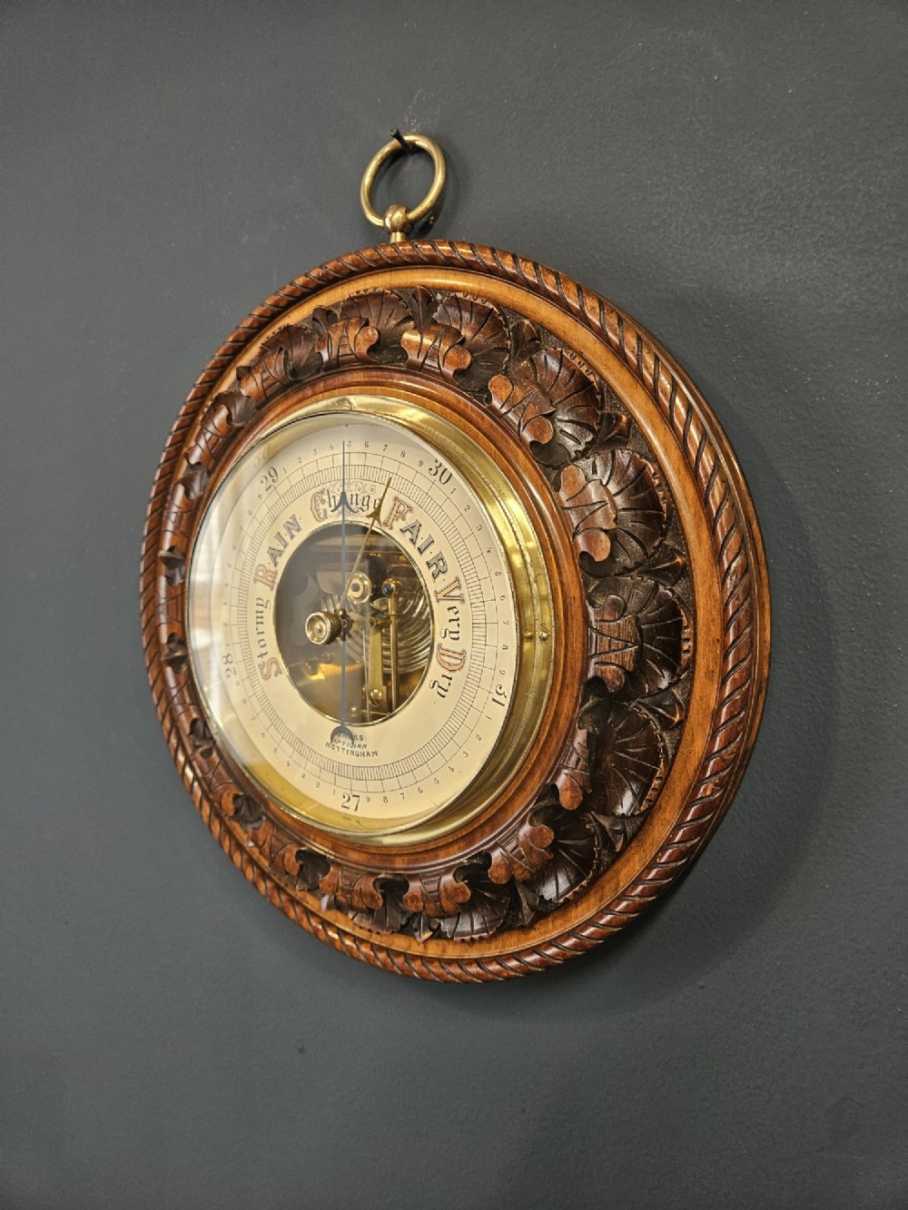 A circular Victorian carved walnut aneroid barometer featuring a rope twist border, leaf motifs, and an open centre dial exposing internal brass mechanisms. View V