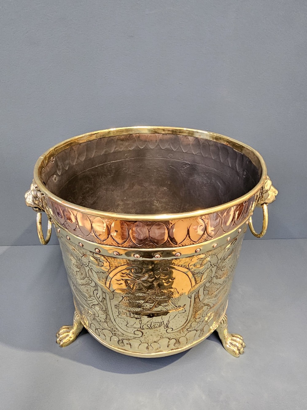 A circular Victorian brass log bin featuring embossed pictorial scenes, a copper rim, cast lion mask side handles, and paw feet.