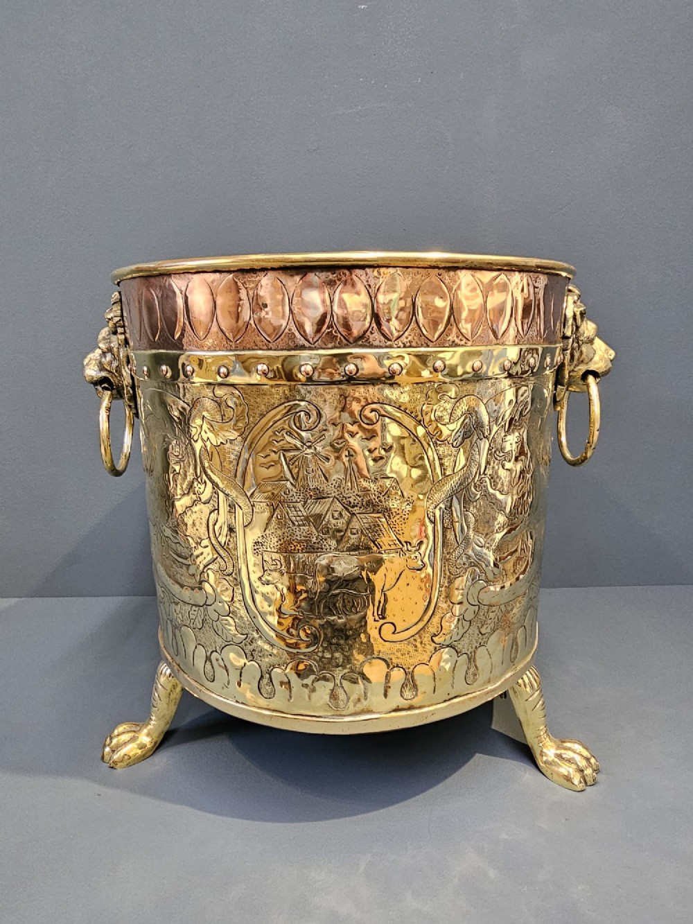 A circular Victorian brass log bin featuring embossed pictorial scenes, a copper rim, cast lion mask side handles, and paw feet. View II