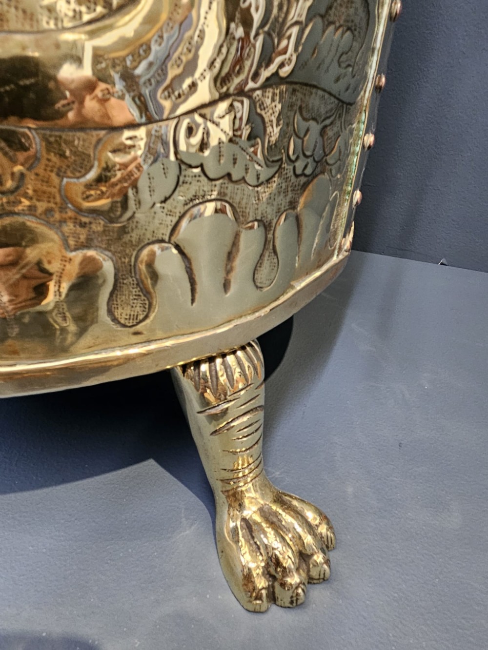 A circular Victorian brass log bin featuring embossed pictorial scenes, a copper rim, cast lion mask side handles, and paw feet. View IV