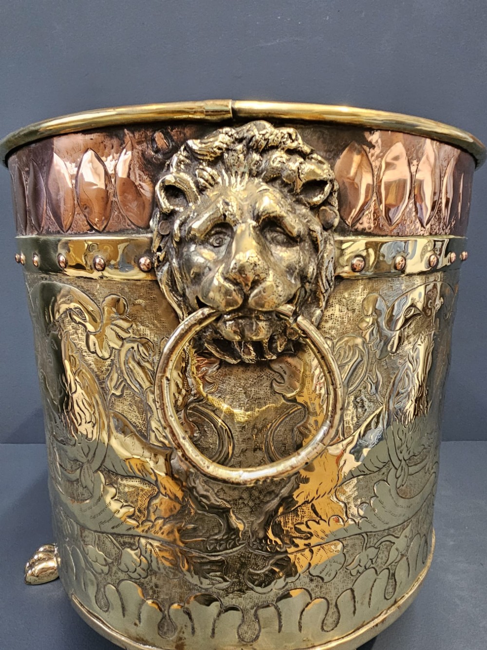 A circular Victorian brass log bin featuring embossed pictorial scenes, a copper rim, cast lion mask side handles, and paw feet. View VI