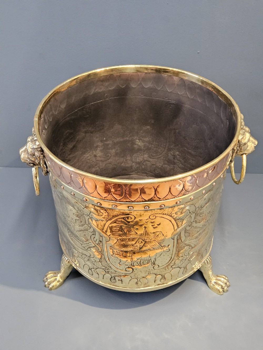 A circular Victorian brass log bin featuring embossed pictorial scenes, a copper rim, cast lion mask side handles, and paw feet. View VIII