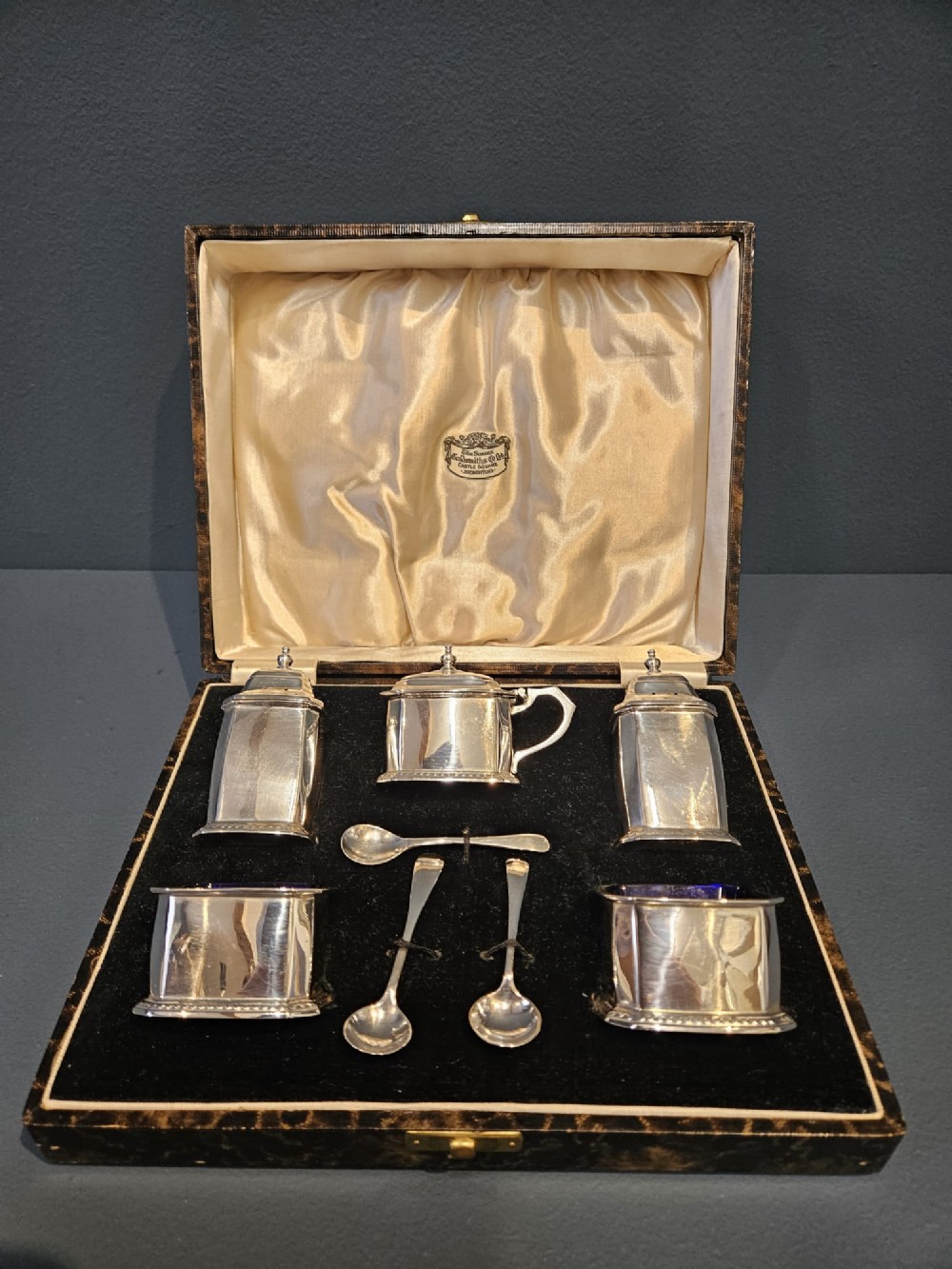 A cased five-piece silver cruet set featuring two pepper casters, two open salt cellars with blue glass liners, a mustard pot, and three spoons inside a silk-lined box.