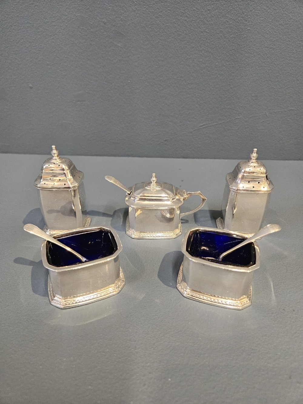 A cased five-piece silver cruet set featuring two pepper casters, two open salt cellars with blue glass liners, a mustard pot, and three spoons inside a silk-lined box. View II