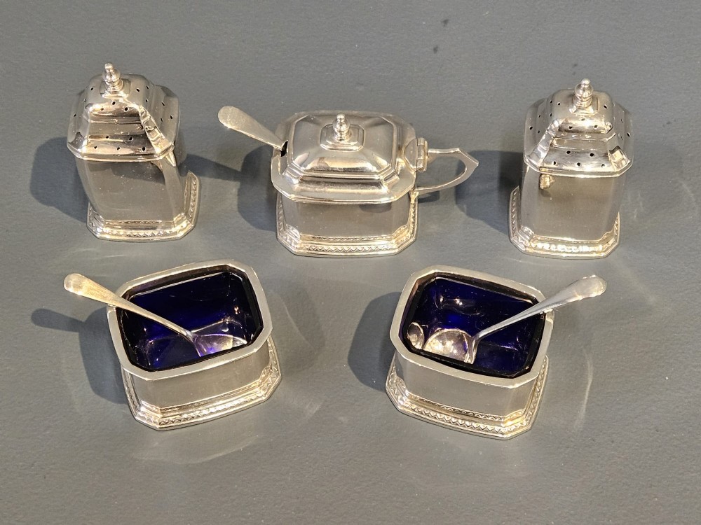 A cased five-piece silver cruet set featuring two pepper casters, two open salt cellars with blue glass liners, a mustard pot, and three spoons inside a silk-lined box. View IV
