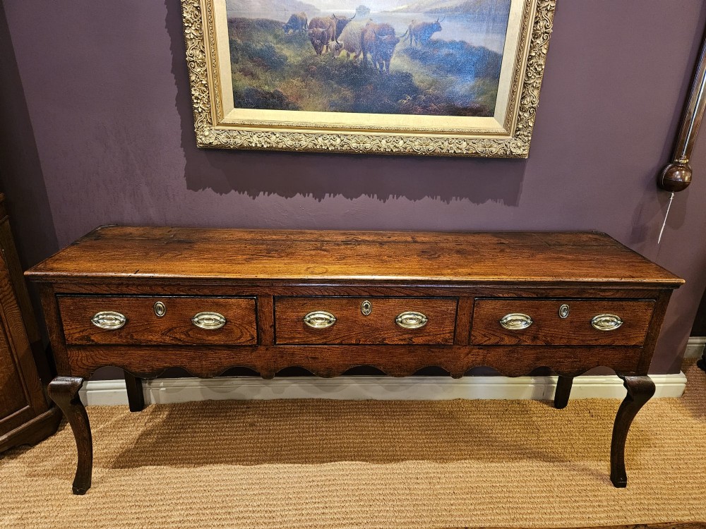 An 18th-century George II elm low dresser featuring three drawers with brass bail handles and shaped cabriole legs.