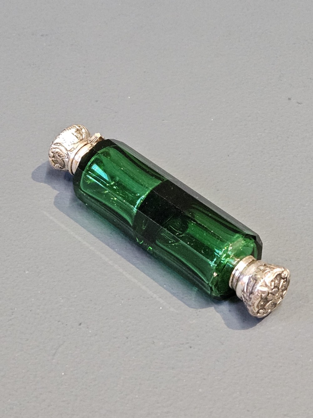 A Victorian double-ended emerald green facet-cut glass scent bottle with detailed embossed silver caps on both ends.