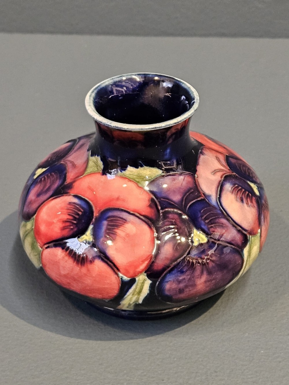 A small squat earthenware vase featuring a dark cobalt blue glaze decorated with hand-painted purple and pink pansy motifs. View II