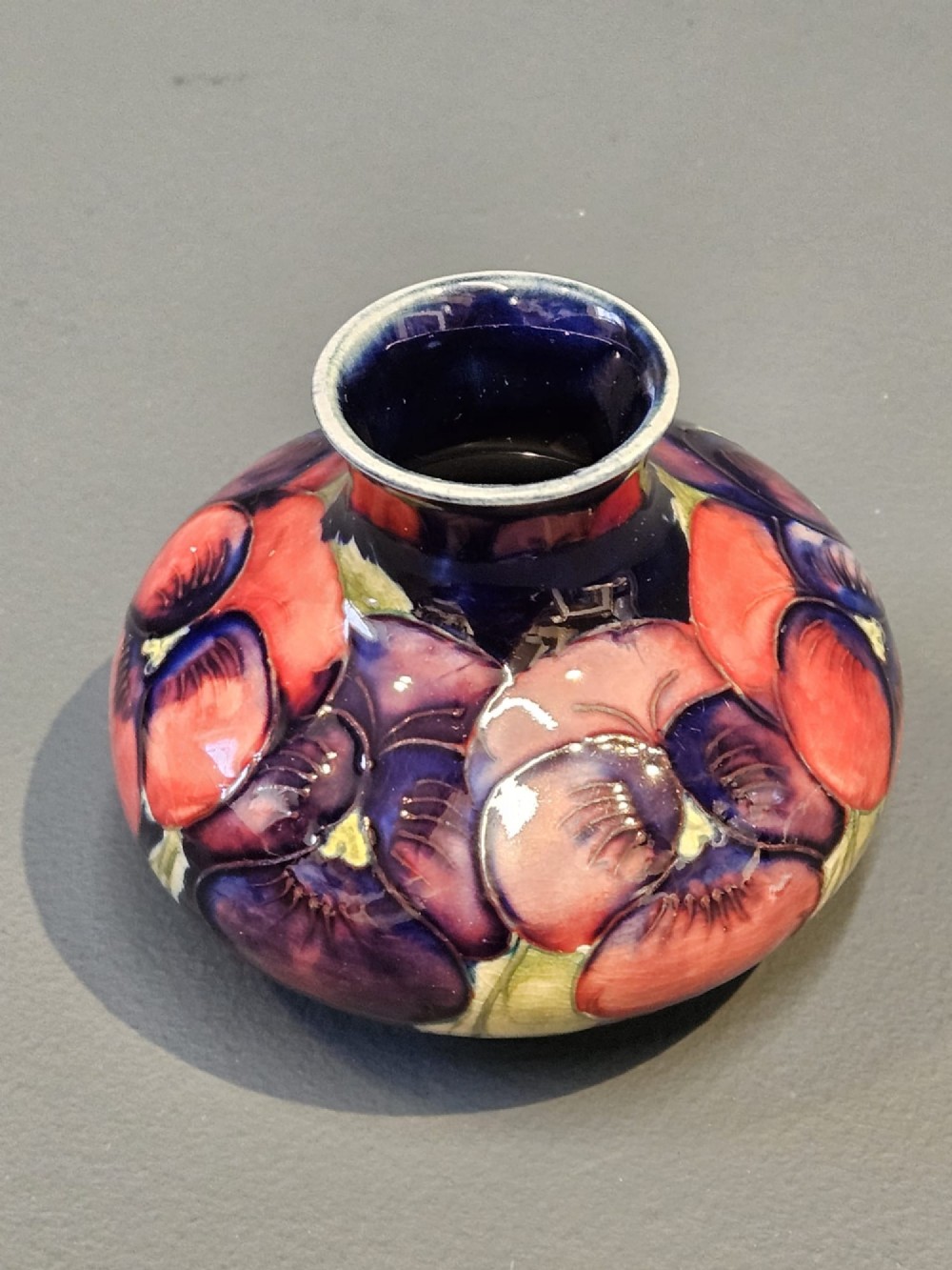 A small squat earthenware vase featuring a dark cobalt blue glaze decorated with hand-painted purple and pink pansy motifs. View III