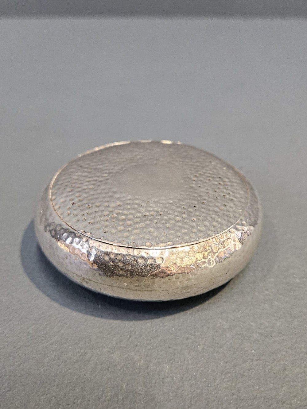 A round, pebble-shaped sterling silver box with a hand-hammered planished texture and smooth central circular cartouche.