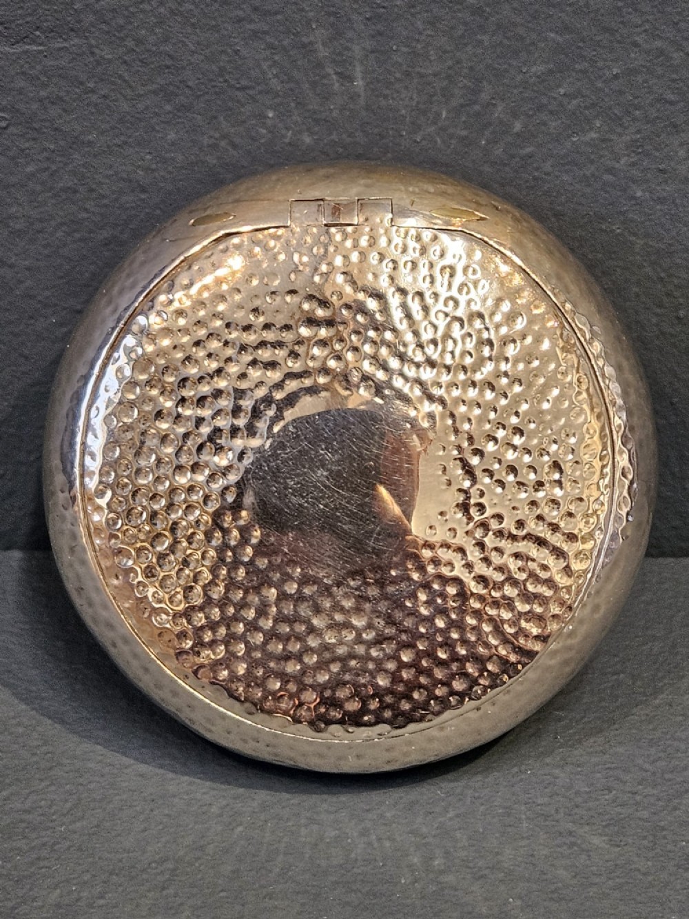 A round, pebble-shaped sterling silver box with a hand-hammered planished texture and smooth central circular cartouche. View VI