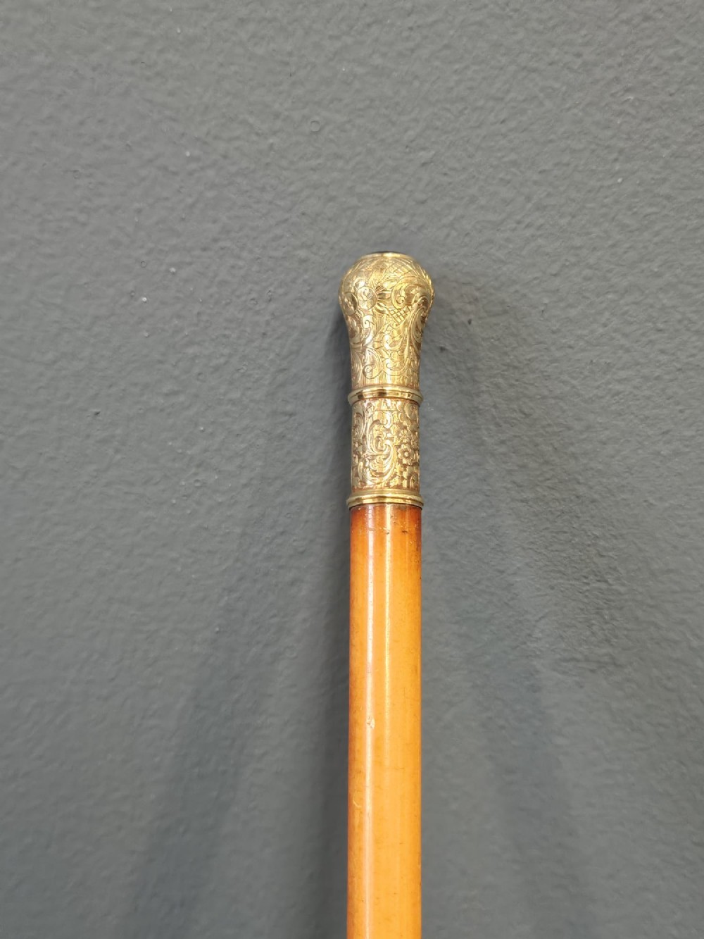 A slender light brown wooden swagger stick featuring a polished gold top inset with a small red ruby. View III