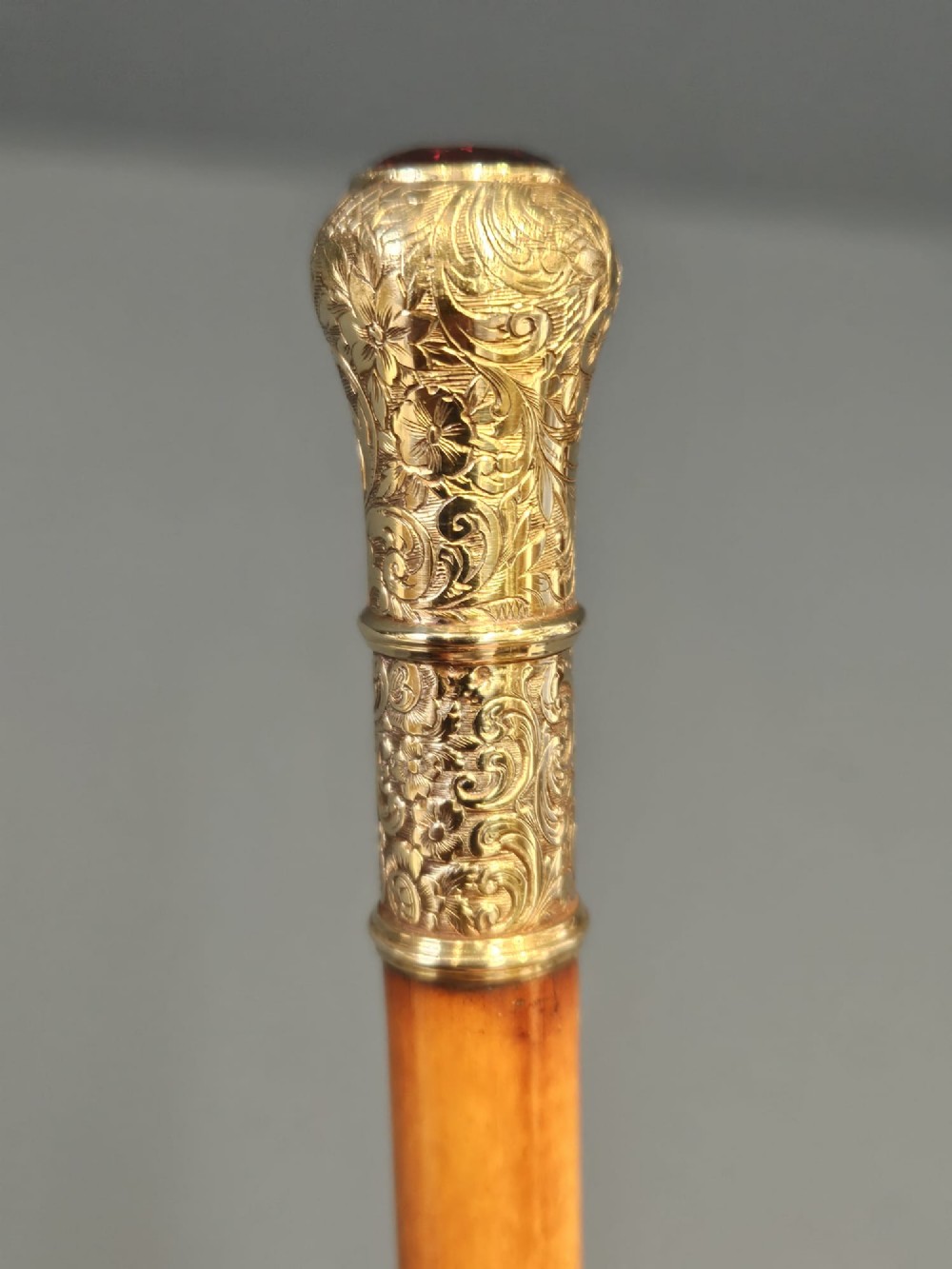 A slender light brown wooden swagger stick featuring a polished gold top inset with a small red ruby. View IV