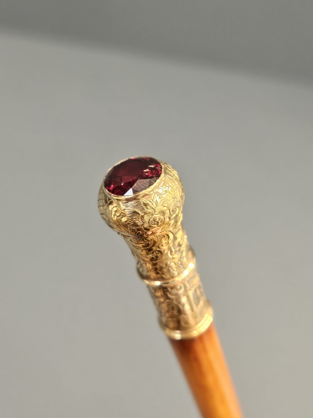 A slender light brown wooden swagger stick featuring a polished gold top inset with a small red ruby. View V