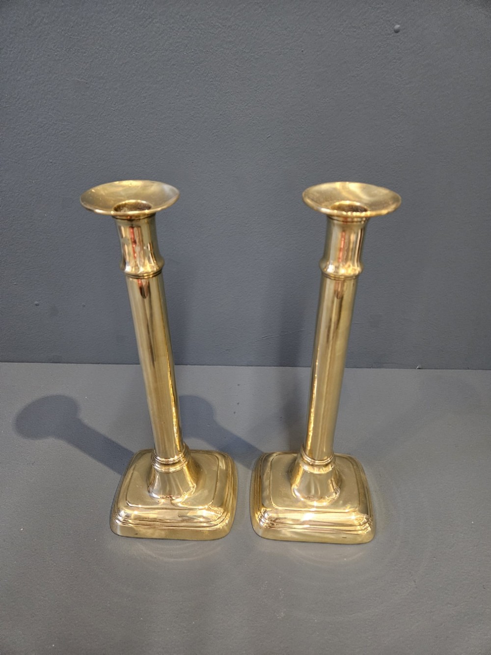 A pair of tall George III brass candlesticks with stepped square bases and slender cylindrical columns featuring raised band details. View II