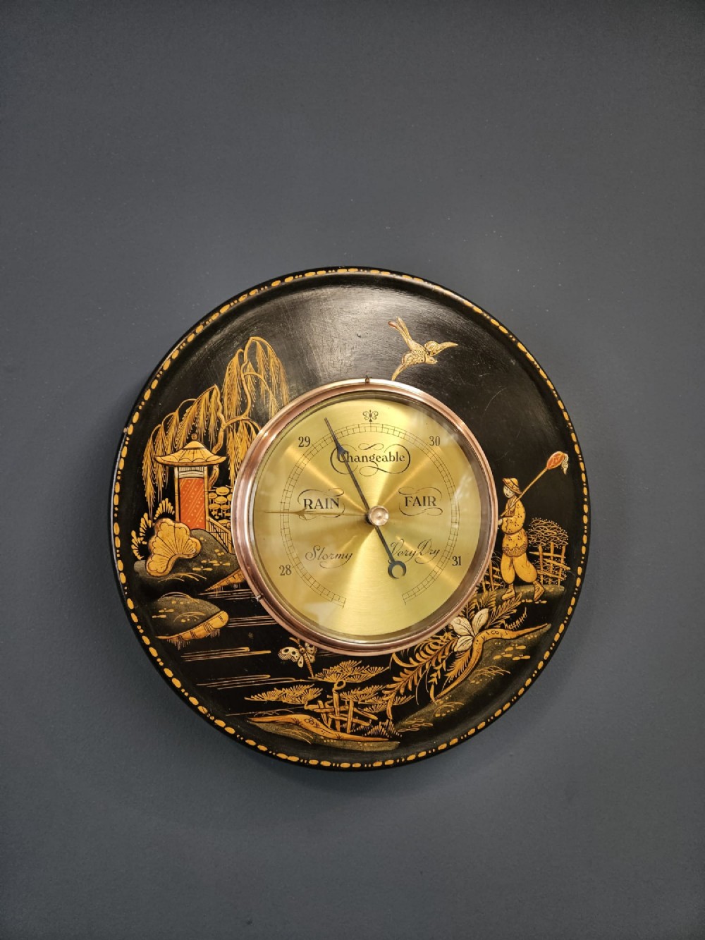 A circular Edwardian aneroid barometer set in a black lacquered frame with gilded hand-painted Chinoiserie figures and landscape motifs.