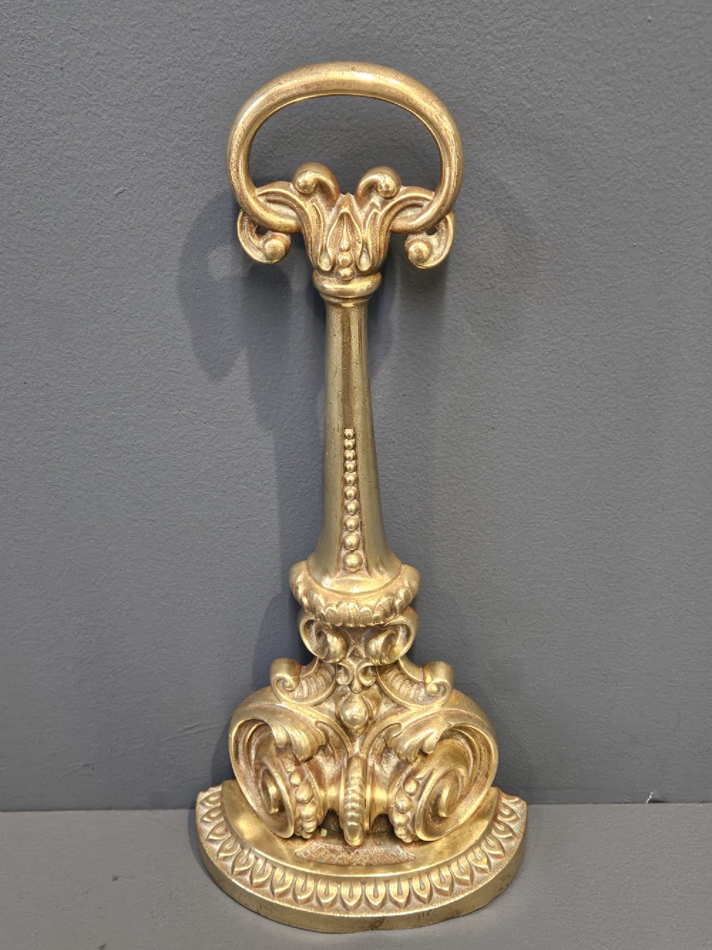 An ornate Victorian cast brass door stop featuring a top ring handle, a tapered column with scrollwork, and a weighted semi-circular base. View III