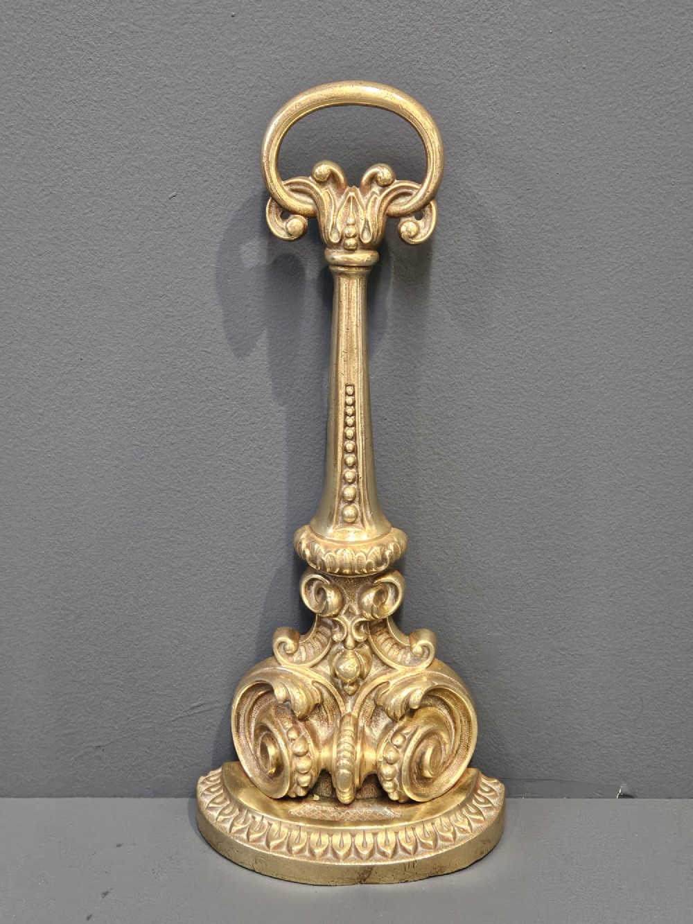 An ornate Victorian cast brass door stop featuring a top ring handle, a tapered column with scrollwork, and a weighted semi-circular base. View IV