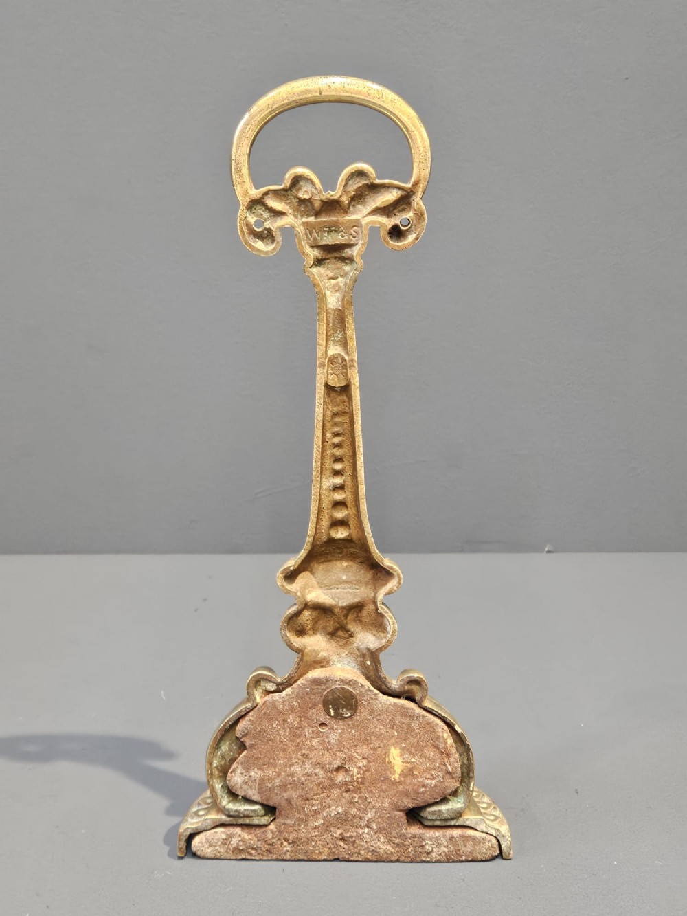 An ornate Victorian cast brass door stop featuring a top ring handle, a tapered column with scrollwork, and a weighted semi-circular base. View V