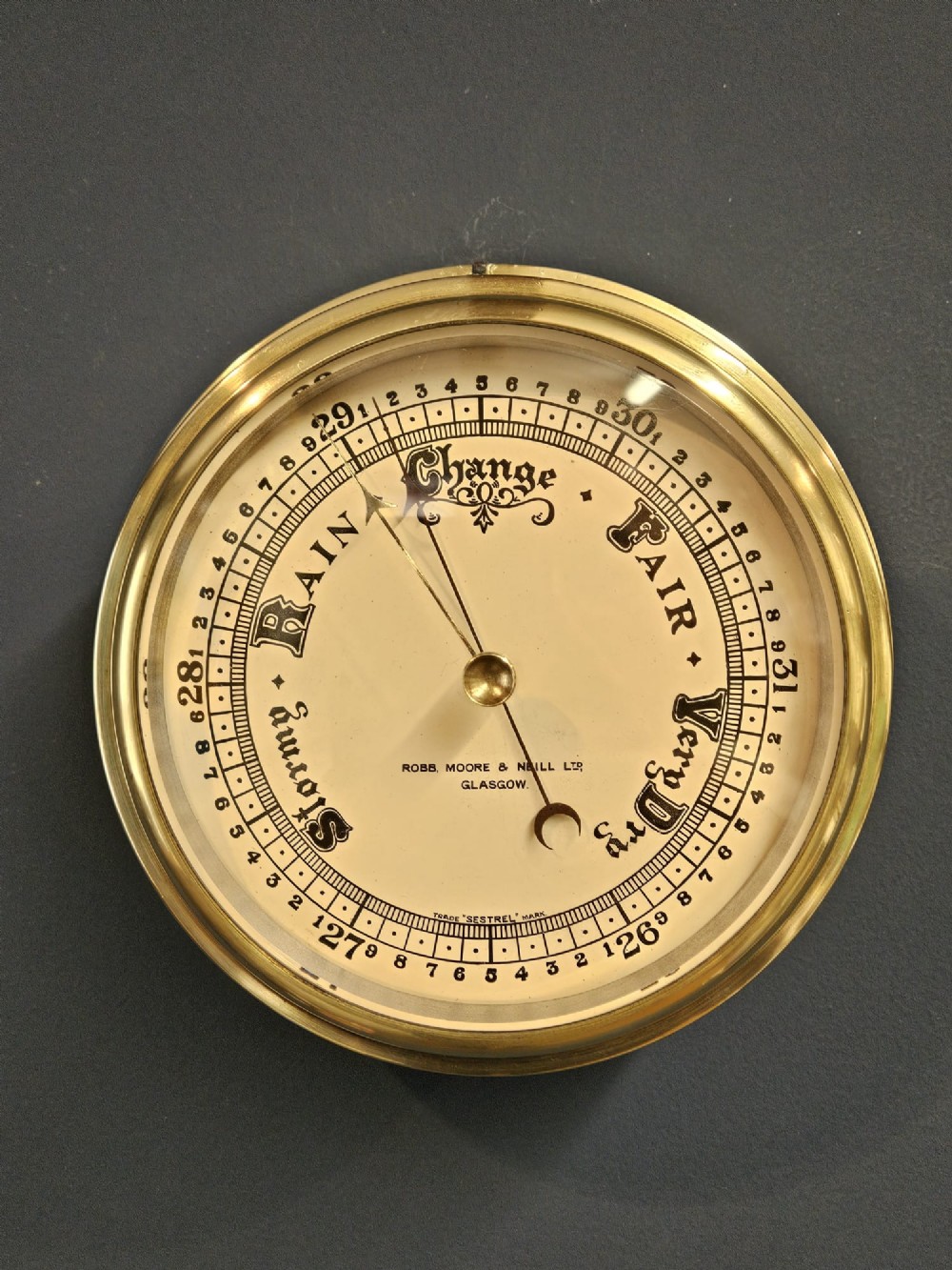 An Edwardian circular brass bulkhead barometer featuring a cream dial with black scale markings, typography, and two pointer hands under a clear glass cover.