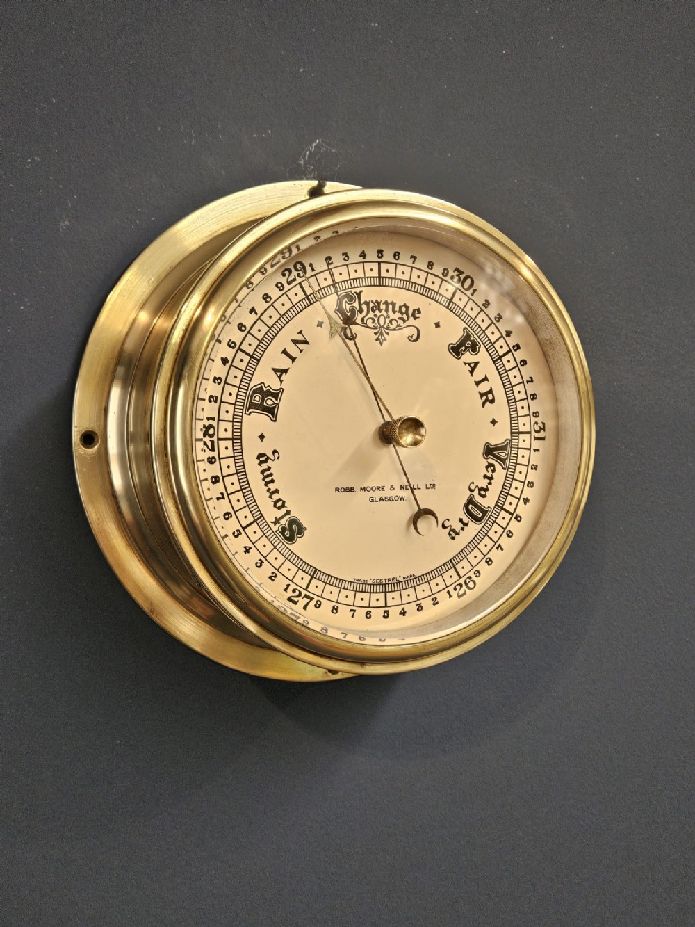 An Edwardian circular brass bulkhead barometer featuring a cream dial with black scale markings, typography, and two pointer hands under a clear glass cover. View IV