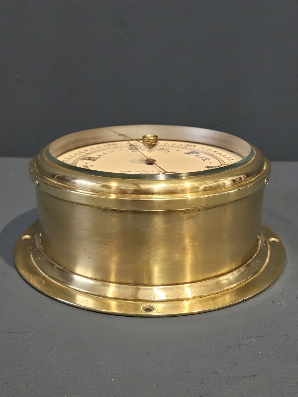 An Edwardian circular brass bulkhead barometer featuring a cream dial with black scale markings, typography, and two pointer hands under a clear glass cover. View V