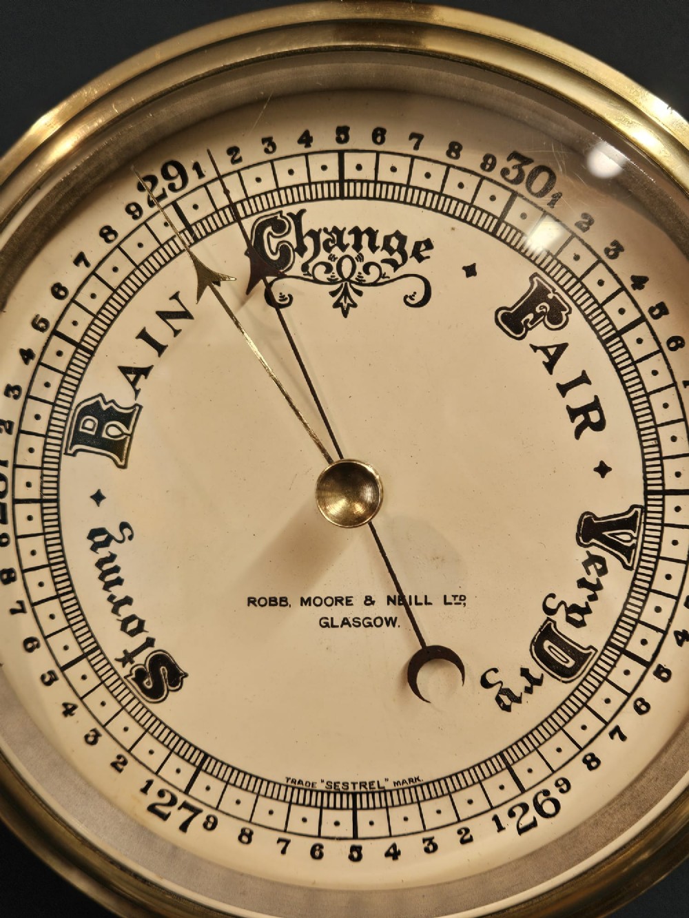 An Edwardian circular brass bulkhead barometer featuring a cream dial with black scale markings, typography, and two pointer hands under a clear glass cover. View VI