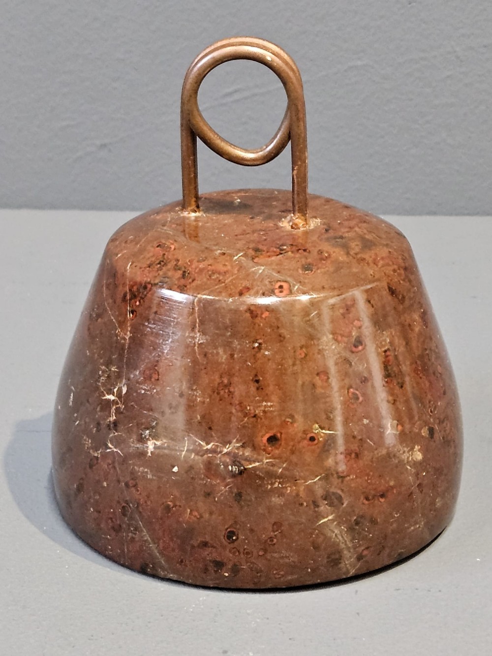 A turned bell-shaped red brown mottled serpentine stone door stop featuring a metal looped wire handle at the top.