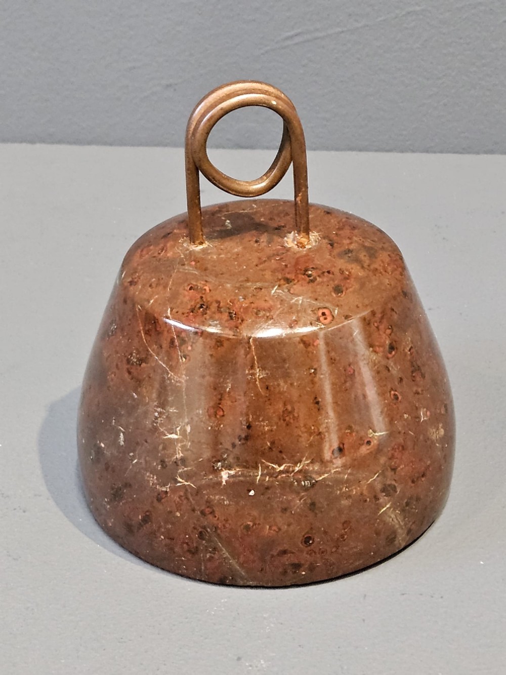A turned bell-shaped red brown mottled serpentine stone door stop featuring a metal looped wire handle at the top. View II