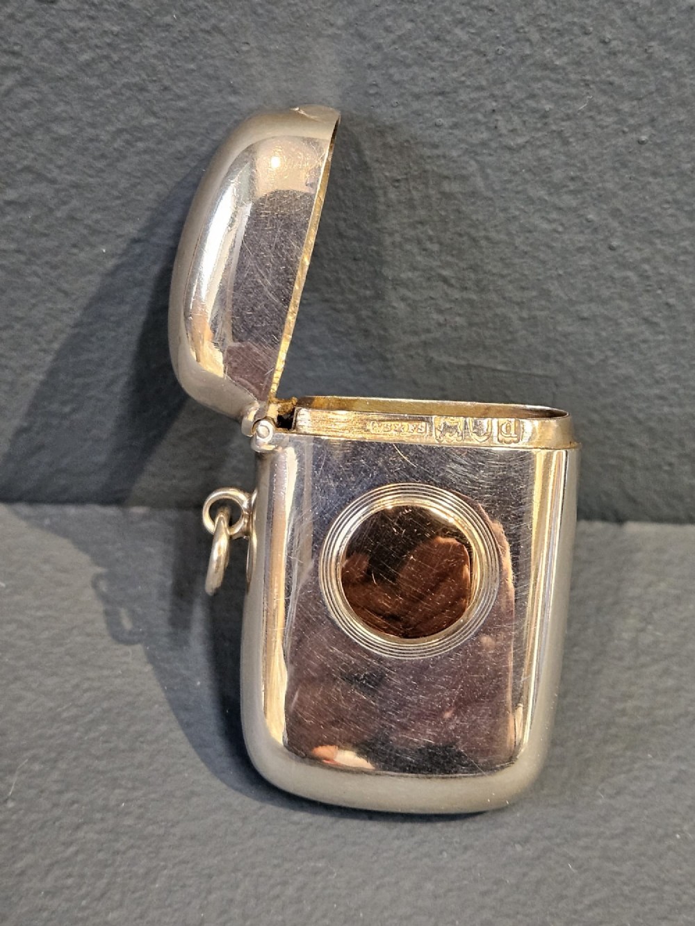 A polished silver vesta case featuring a central circular gold plaque and a side ring attachment. View II