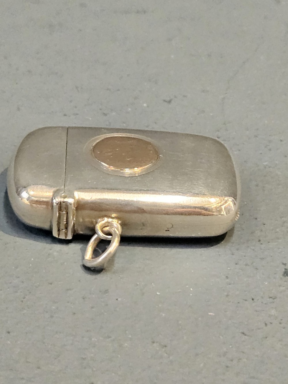 A polished silver vesta case featuring a central circular gold plaque and a side ring attachment. View III
