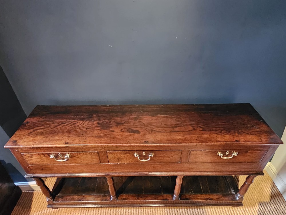An 18th-century George II oak pot board dresser featuring three frieze drawers with brass handles set above an open lower shelf and turned baluster supports. View V