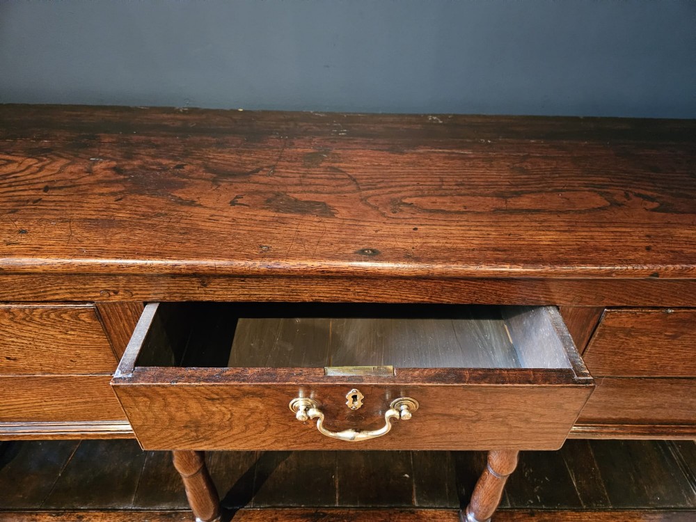 An 18th-century George II oak pot board dresser featuring three frieze drawers with brass handles set above an open lower shelf and turned baluster supports. View IX