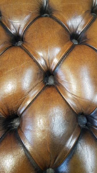 A 1930s tan leather footstool featuring an inclined top with deep buttoning, brass stud trim, and dark wooden legs. View V