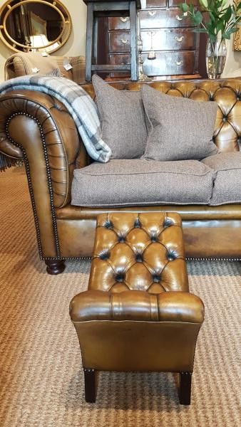 A 1930s tan leather footstool featuring an inclined top with deep buttoning, brass stud trim, and dark wooden legs. View VI
