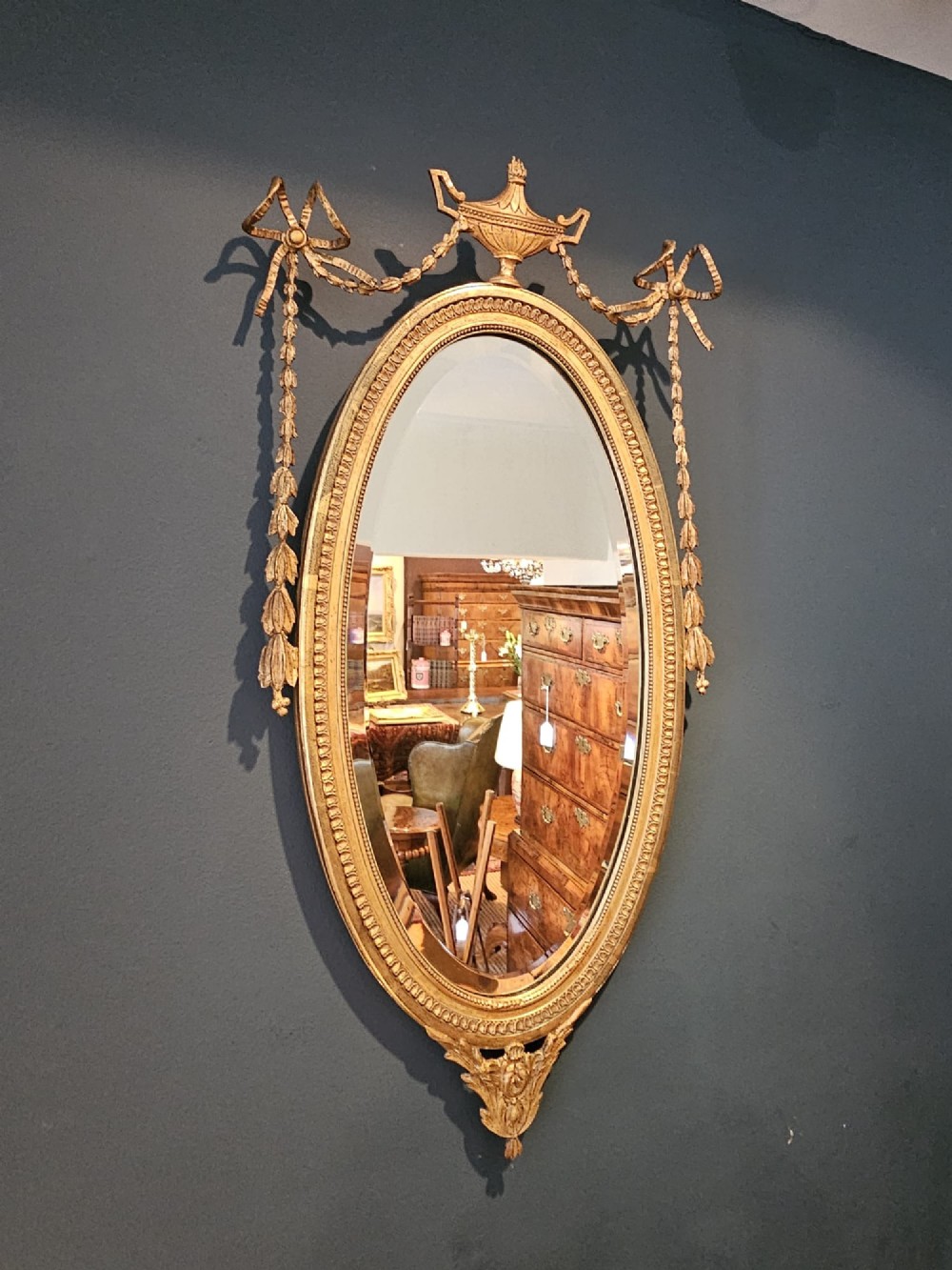An oval giltwood wall mirror featuring a carved urn pediment with draped ribbon swags, a beaded inner sight edge, and a decorative lower apron. View II
