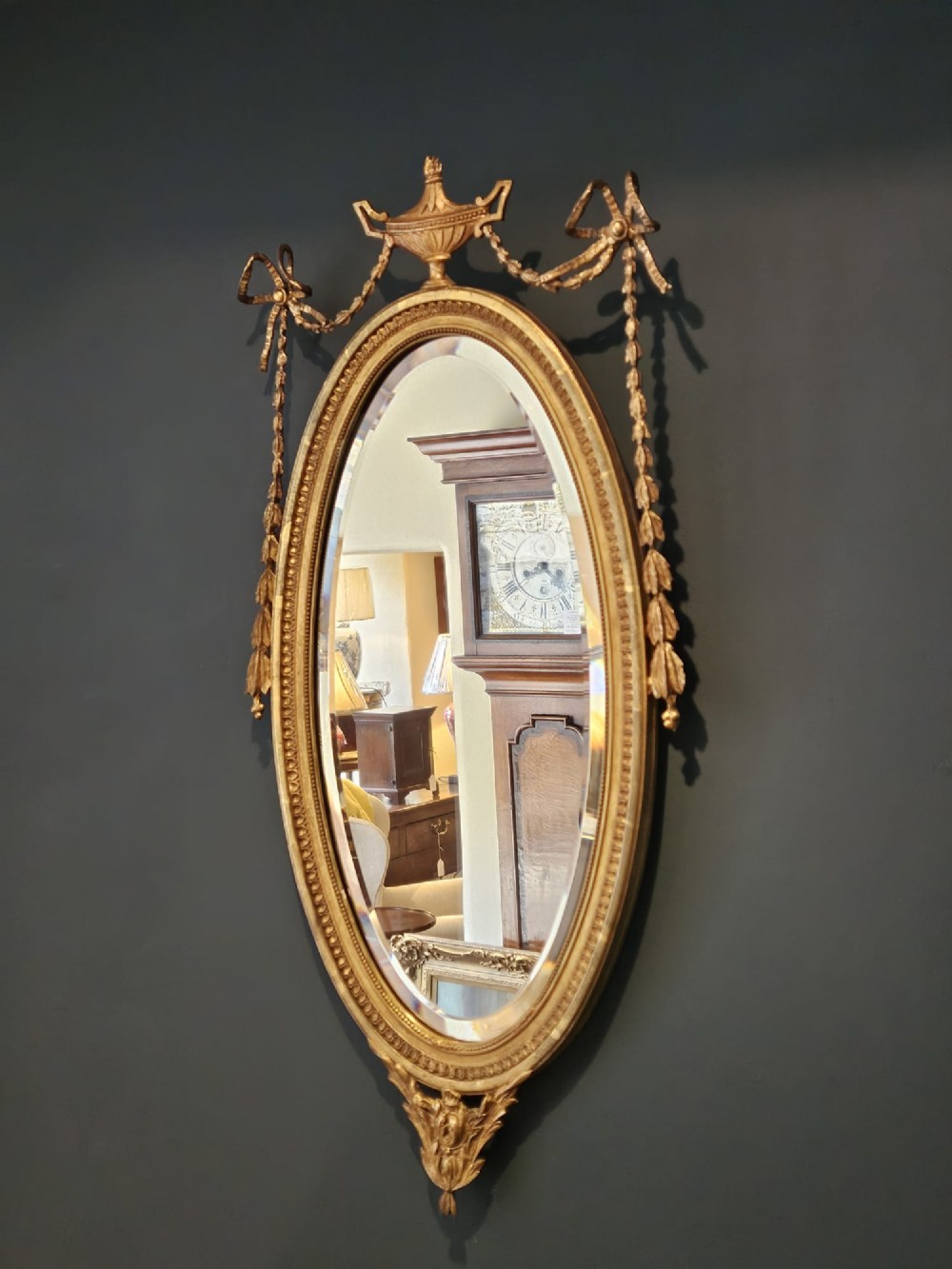 An oval giltwood wall mirror featuring a carved urn pediment with draped ribbon swags, a beaded inner sight edge, and a decorative lower apron. View III