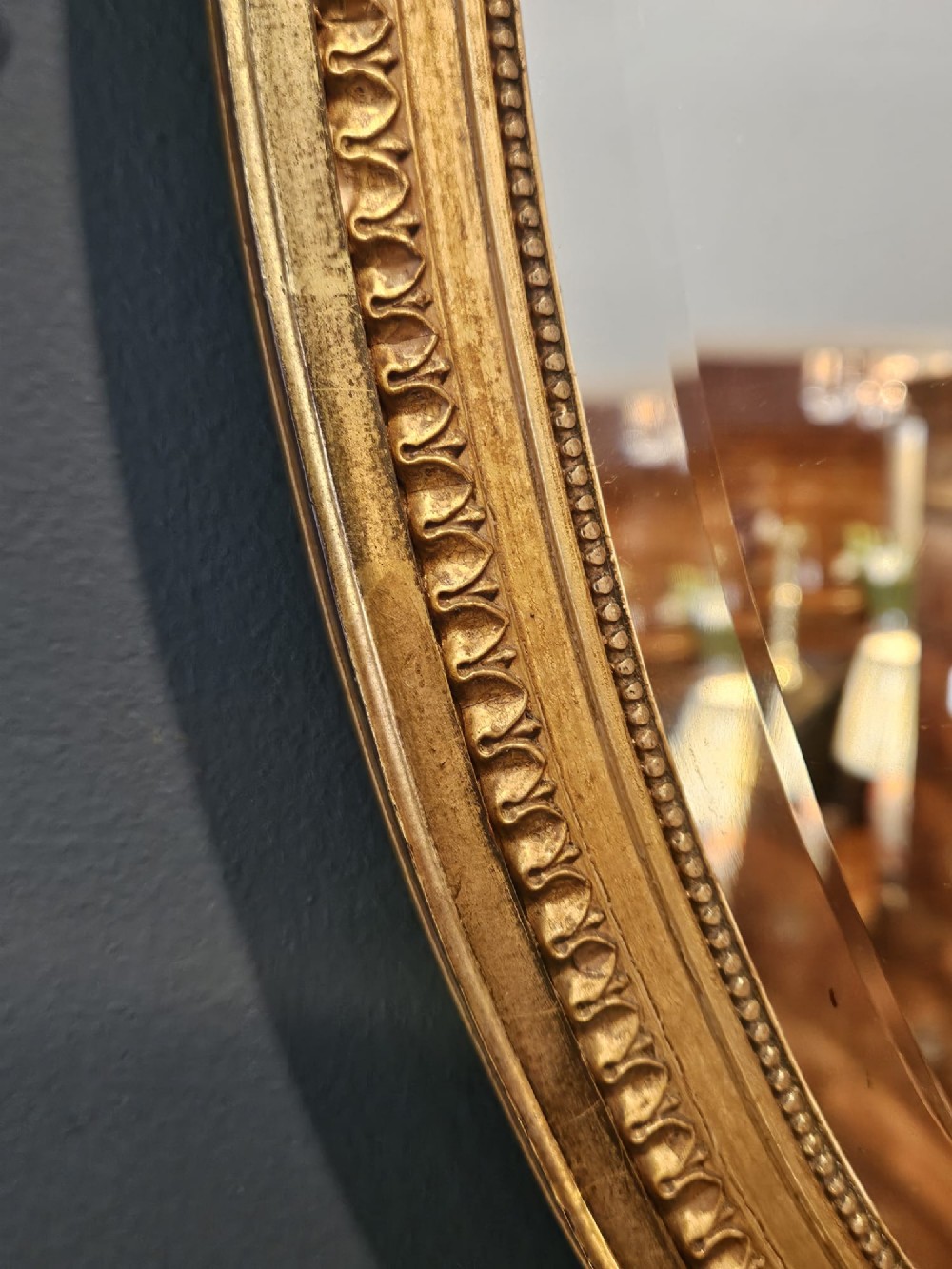 An oval giltwood wall mirror featuring a carved urn pediment with draped ribbon swags, a beaded inner sight edge, and a decorative lower apron. View IV