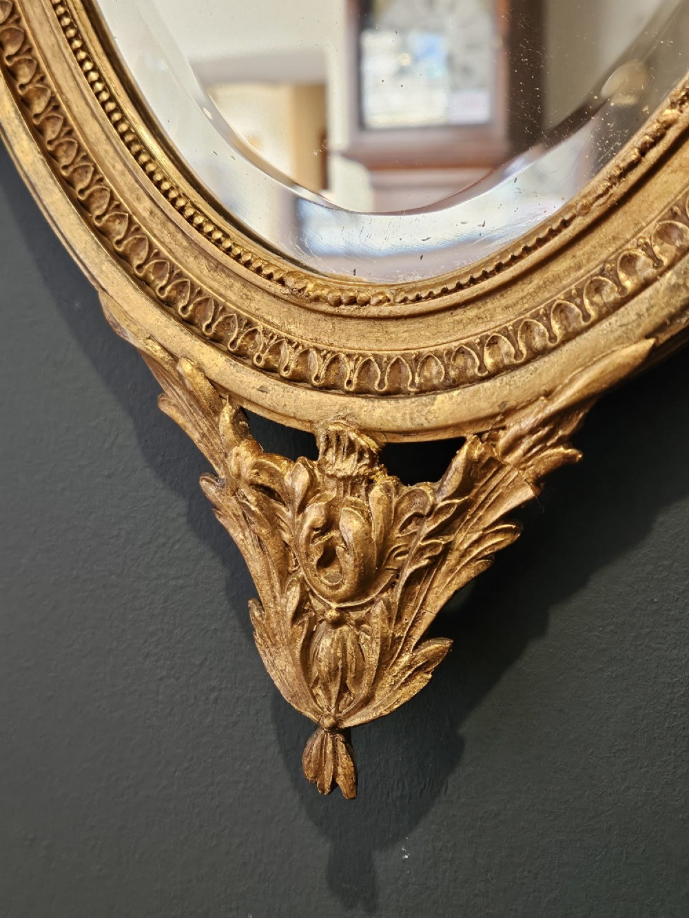 An oval giltwood wall mirror featuring a carved urn pediment with draped ribbon swags, a beaded inner sight edge, and a decorative lower apron. View V