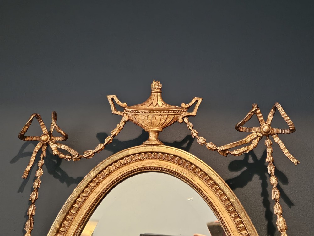 An oval giltwood wall mirror featuring a carved urn pediment with draped ribbon swags, a beaded inner sight edge, and a decorative lower apron. View VI