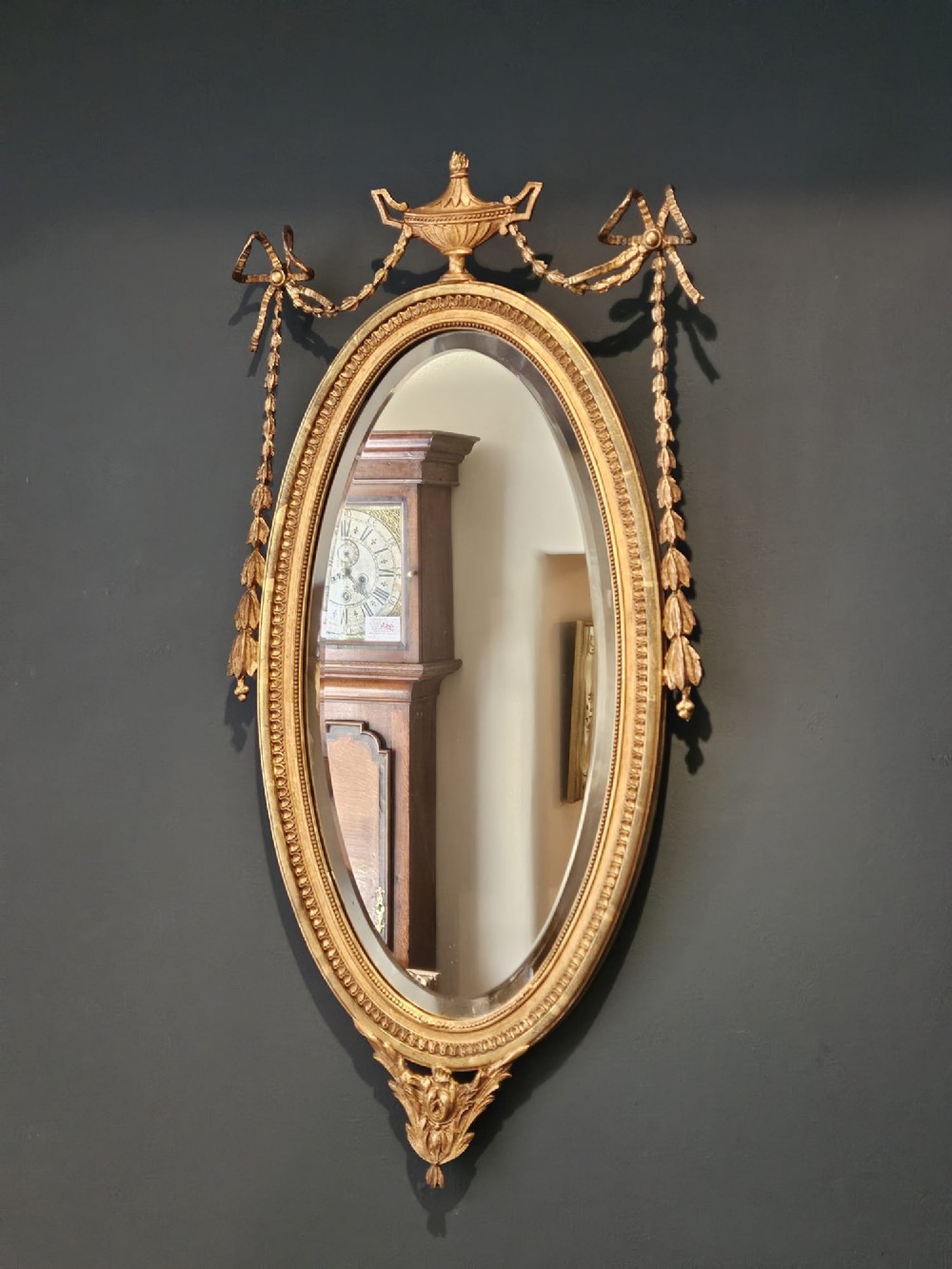 An oval giltwood wall mirror featuring a carved urn pediment with draped ribbon swags, a beaded inner sight edge, and a decorative lower apron. View VII