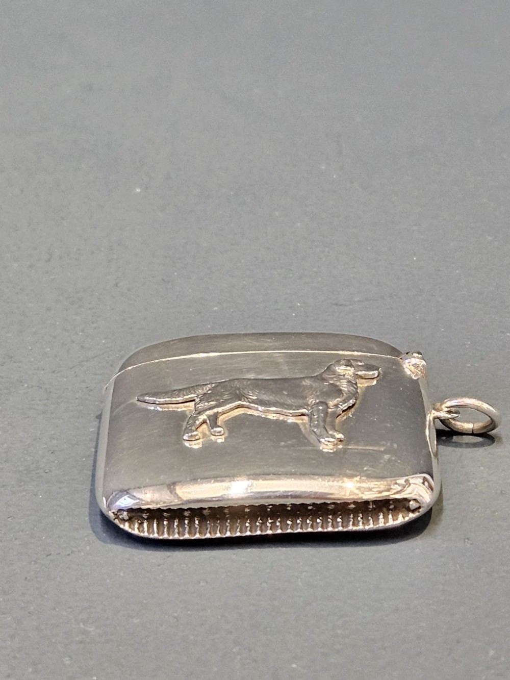 A polished silver rectangular vesta case with a hinged lid, side loop, and an embossed retriever dog motif in relief. View II
