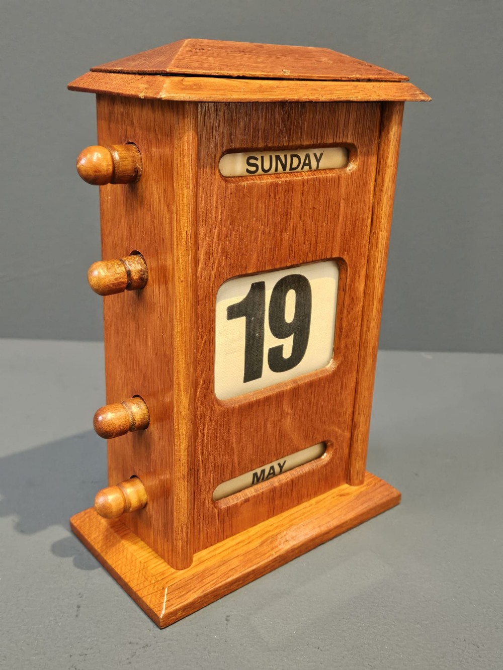 A golden oak perpetual desk calendar featuring side turning knobs and printed aperture windows displaying the day, date, and month. View III