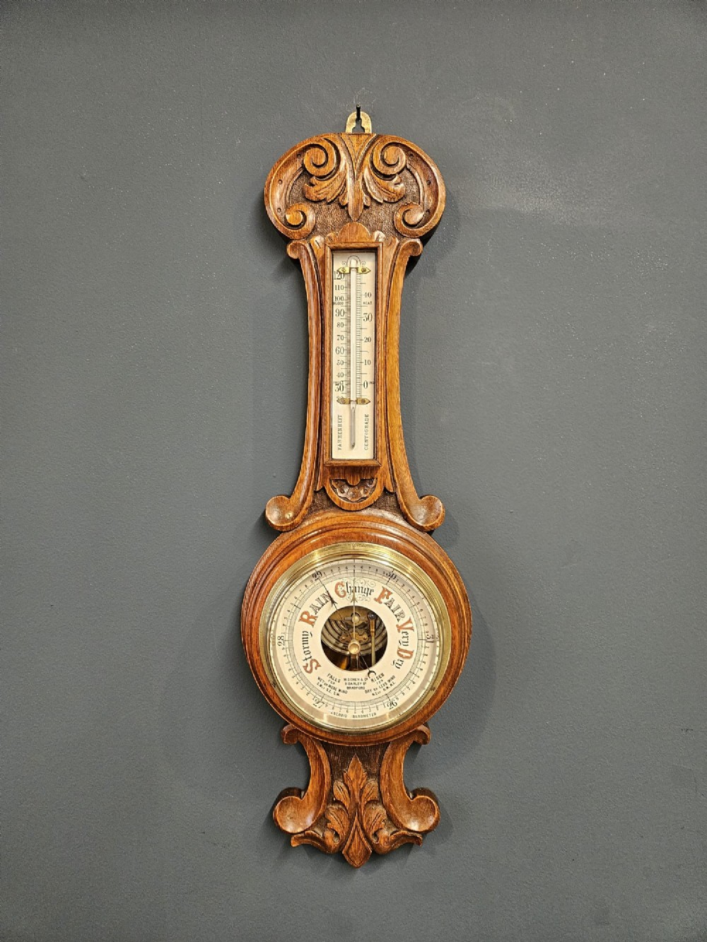 A carved oak banjo barometer featuring a upper spirit thermometer and a circular porcelain dial with an exposed skeletonised movement.