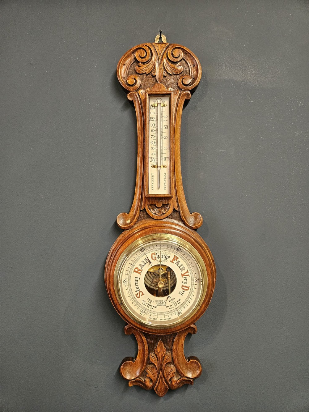 A carved oak banjo barometer featuring a upper spirit thermometer and a circular porcelain dial with an exposed skeletonised movement. View II