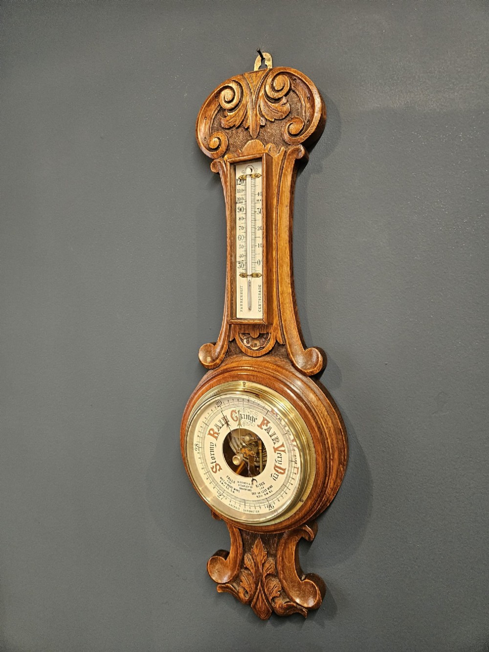 A carved oak banjo barometer featuring a upper spirit thermometer and a circular porcelain dial with an exposed skeletonised movement. View IV