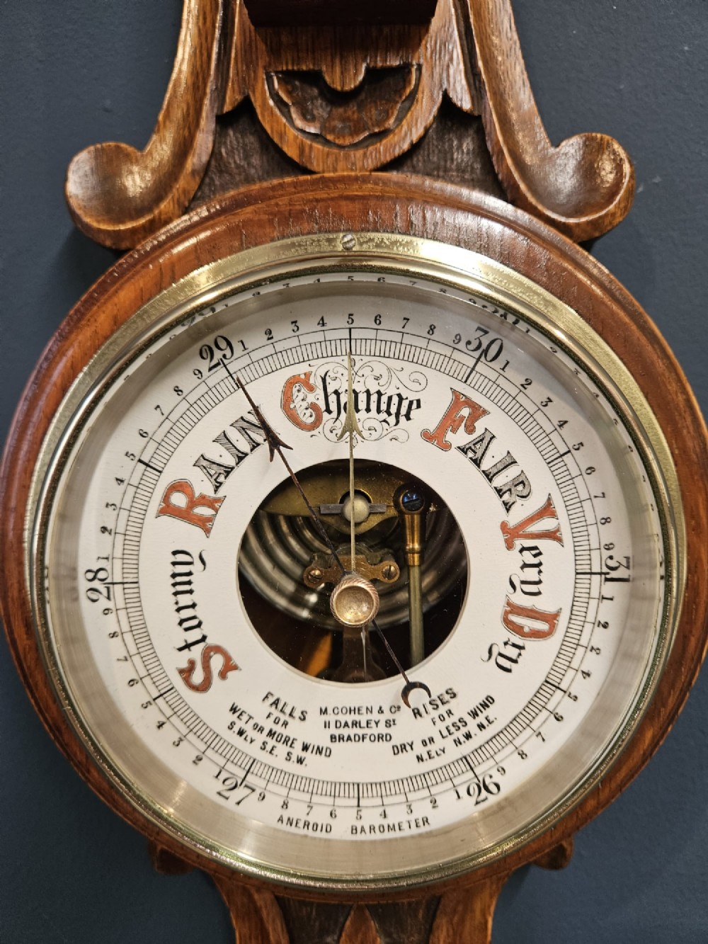 A carved oak banjo barometer featuring a upper spirit thermometer and a circular porcelain dial with an exposed skeletonised movement. View V