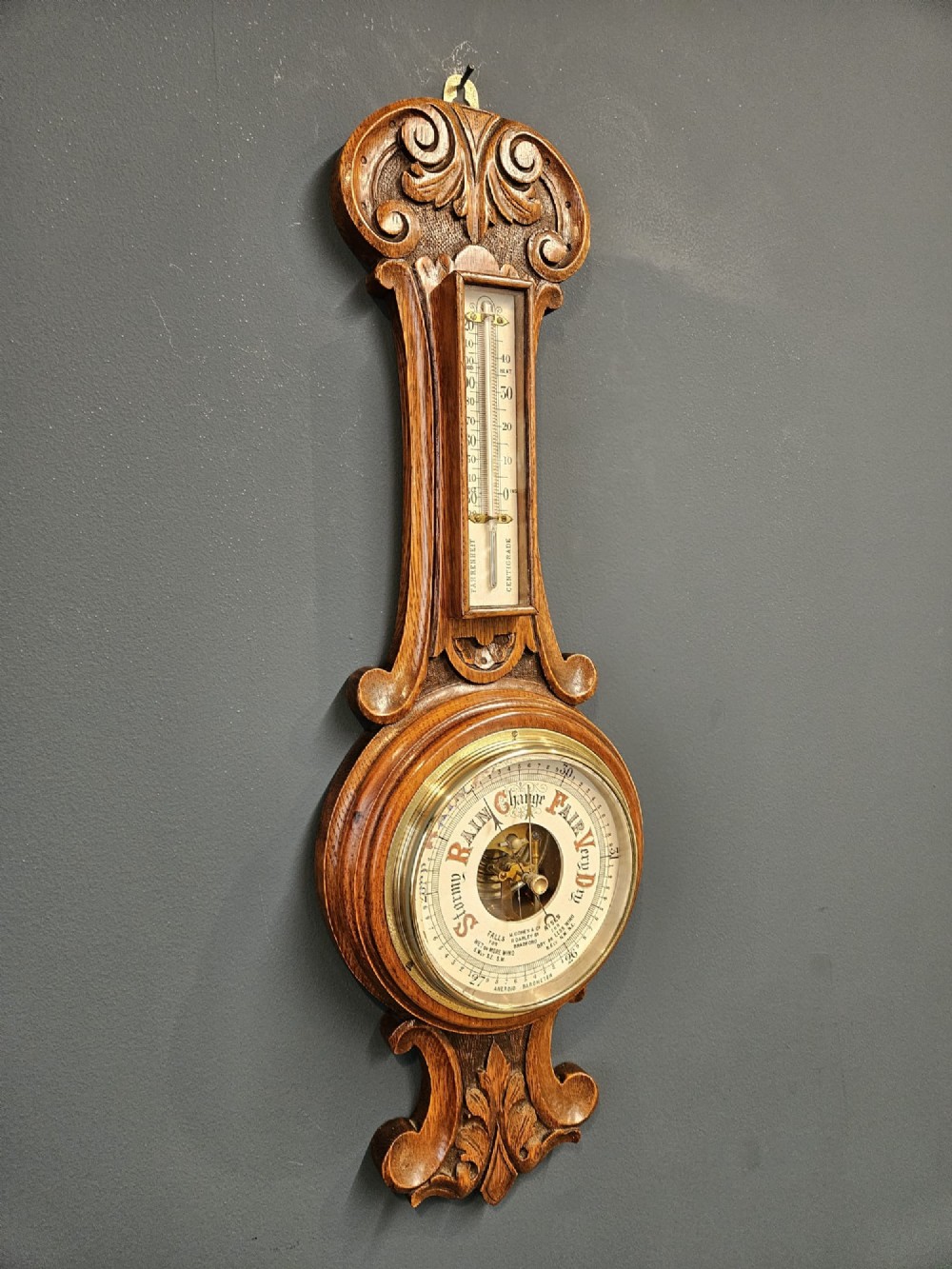 A carved oak banjo barometer featuring a upper spirit thermometer and a circular porcelain dial with an exposed skeletonised movement. View VI
