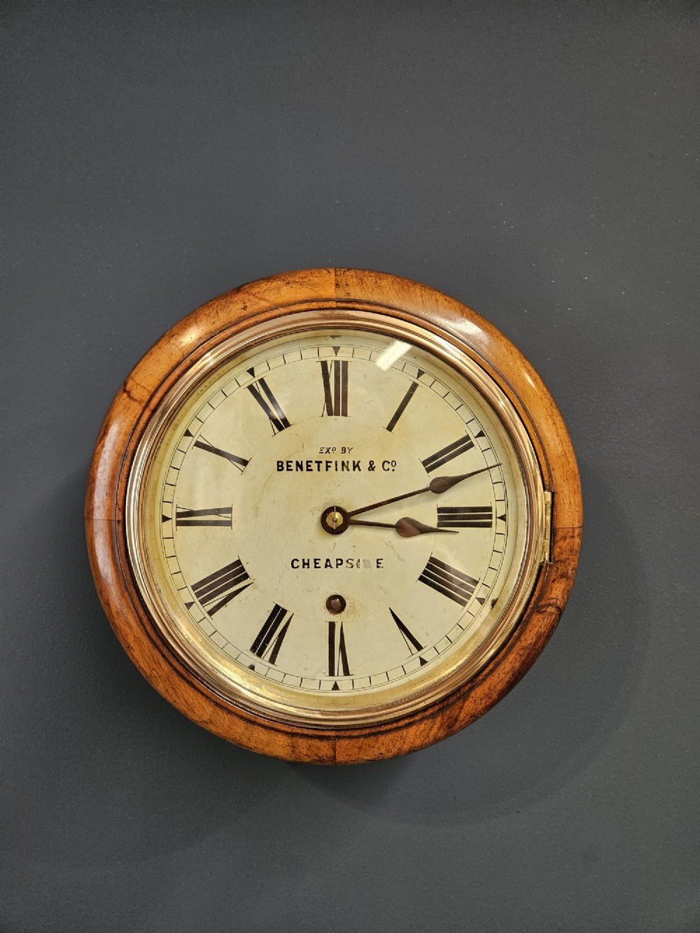 A circular Victorian wall clock featuring a turned warm brown walnut frame, brass-rimmed glass door, and off-white dial marked with Roman numerals and Benetfink and Co Cheapside.