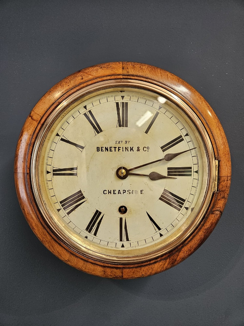 A circular Victorian wall clock featuring a turned warm brown walnut frame, brass-rimmed glass door, and off-white dial marked with Roman numerals and Benetfink and Co Cheapside. View II